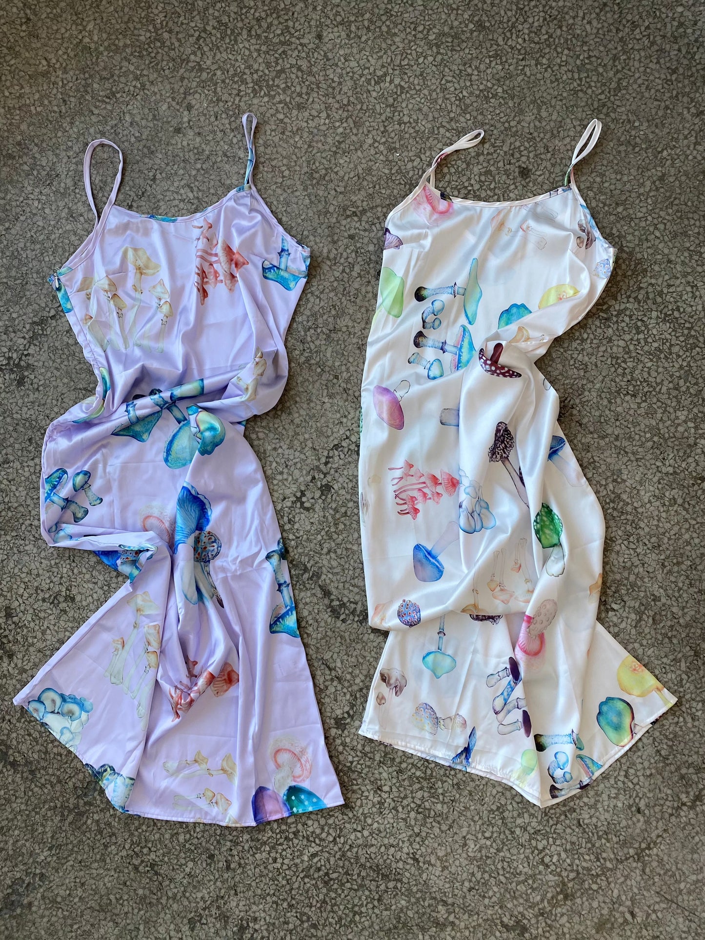 two slip dresses lay crumpled up on the concrete floor, one is lilac with a psychedlic mushroom print and the other has a pink base with a similar mushroom print