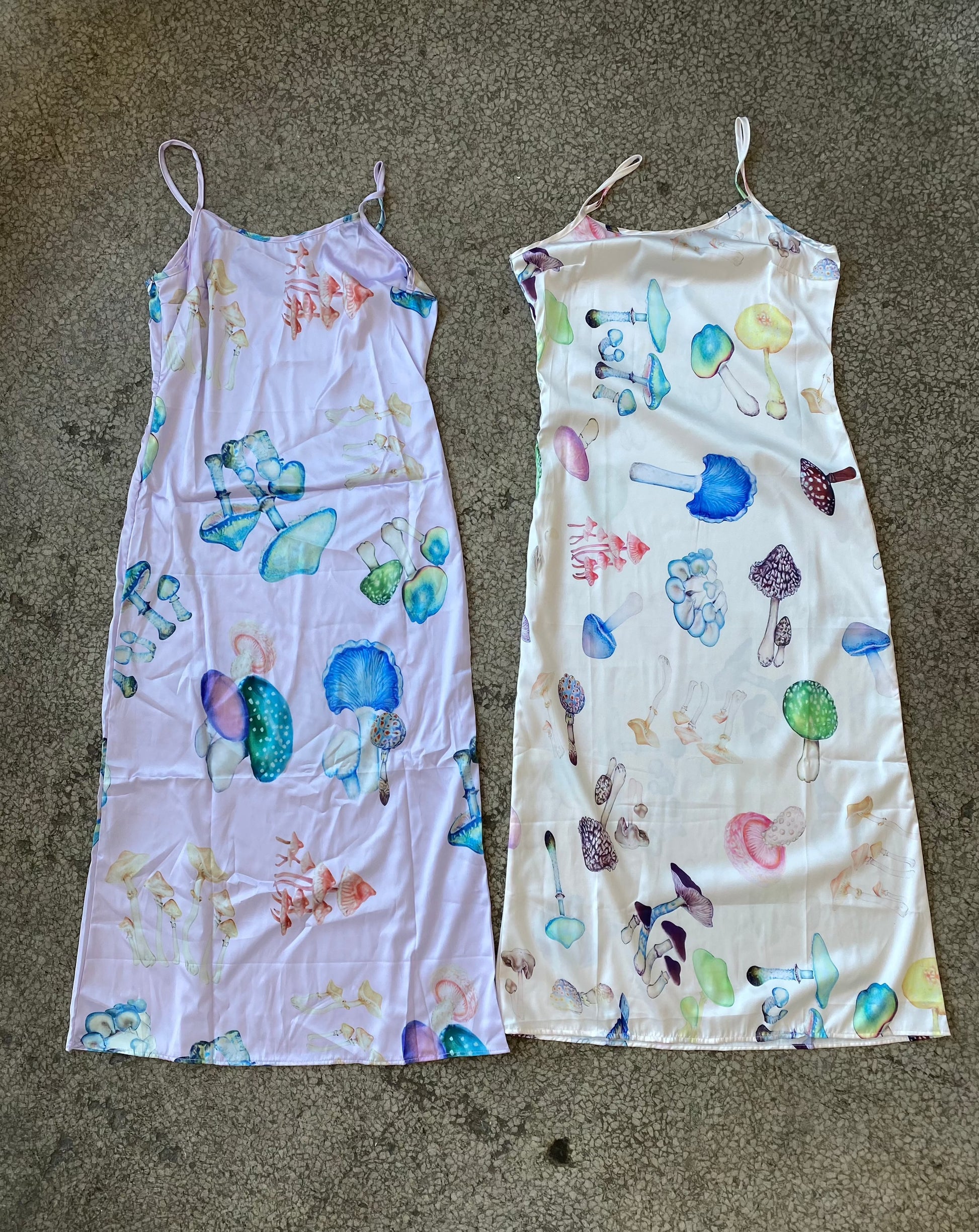 two slip dresses lay side by side on the concrete floor, one is lilac with a psychedlic mushroom print and the other has a pink base with a similar mushroom print
