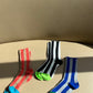 three pairs of socks are sitting on top of a caramel color couch, one pair is red and taupe stripes with a bright blue cap, the middle pair is a vertical black and while stripe sock with a lime green cap and the pair on the right is dodger blue and gray with an emerald green cap