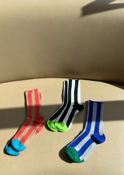 three pairs of socks are sitting on top of a caramel color couch, one pair is red and taupe stripes with a bright blue cap, the middle pair is a vertical black and while stripe sock with a lime green cap and the pair on the right is dodger blue and gray with an emerald green cap