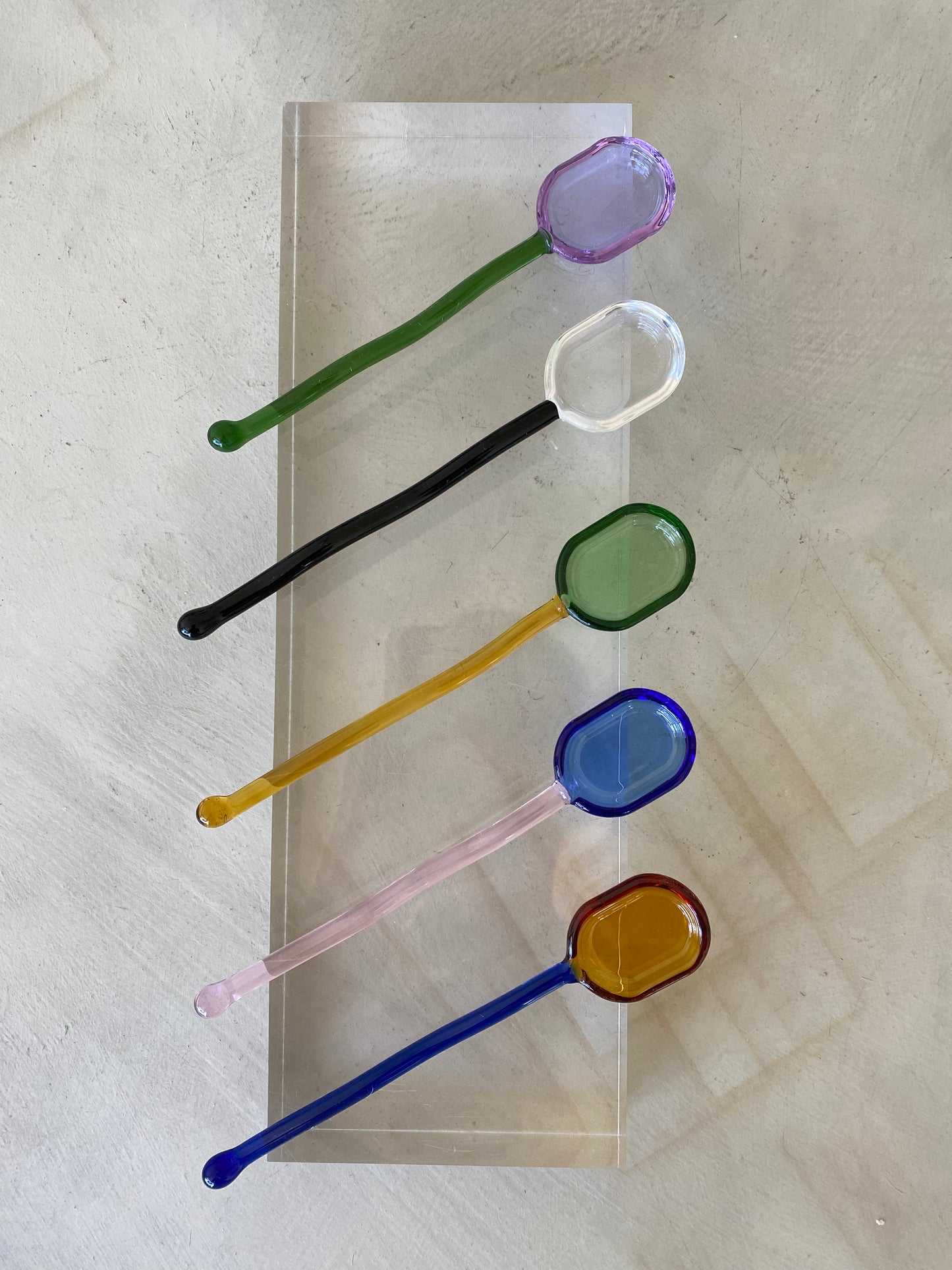 5 glass spoons are arranged on top of a lucite block, from the top to bottom the top spoon has a green handle and a violet bowl, the next one down has a black handle and a clear glass bowl, the middle spoon has a yellow handle and a green bowl, the 4th spoon down from the top is pink with a blue bowl, and the 5th spoon at the bottom of the stack has a cobalt handle and amber bowl